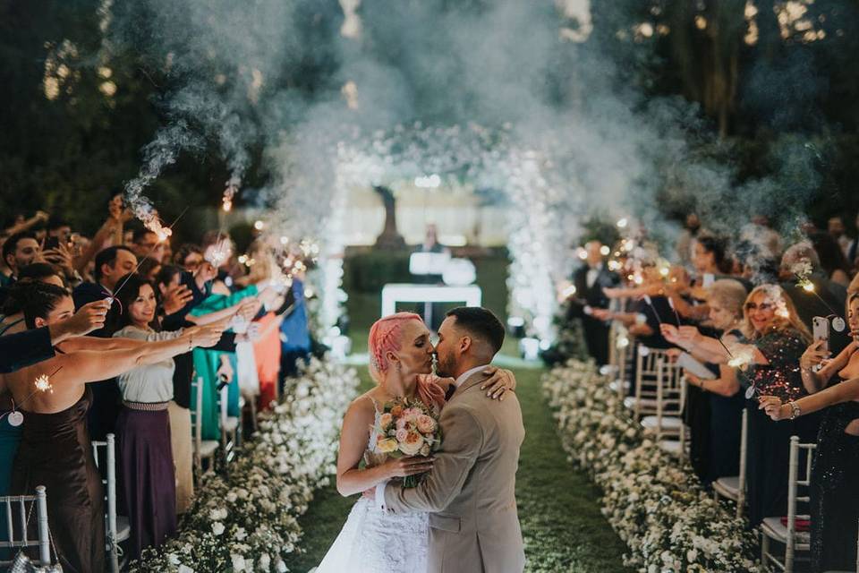 Pareja besándose en un pasillo de flores entre sus invitados